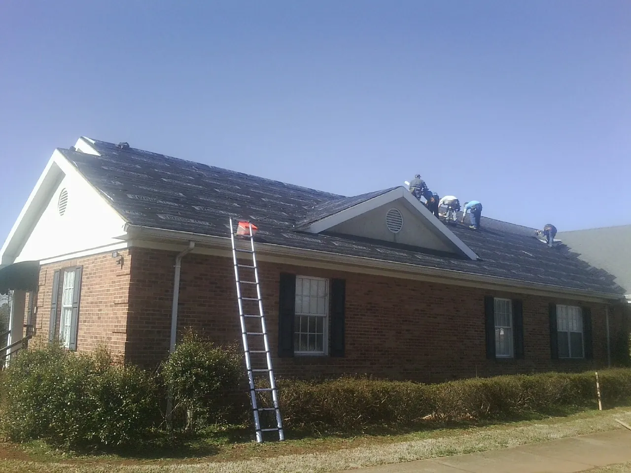 Crew tearing shingles off a brick ranch roof with a ladder set — Emergency Roof Repair work by our crew in Lind