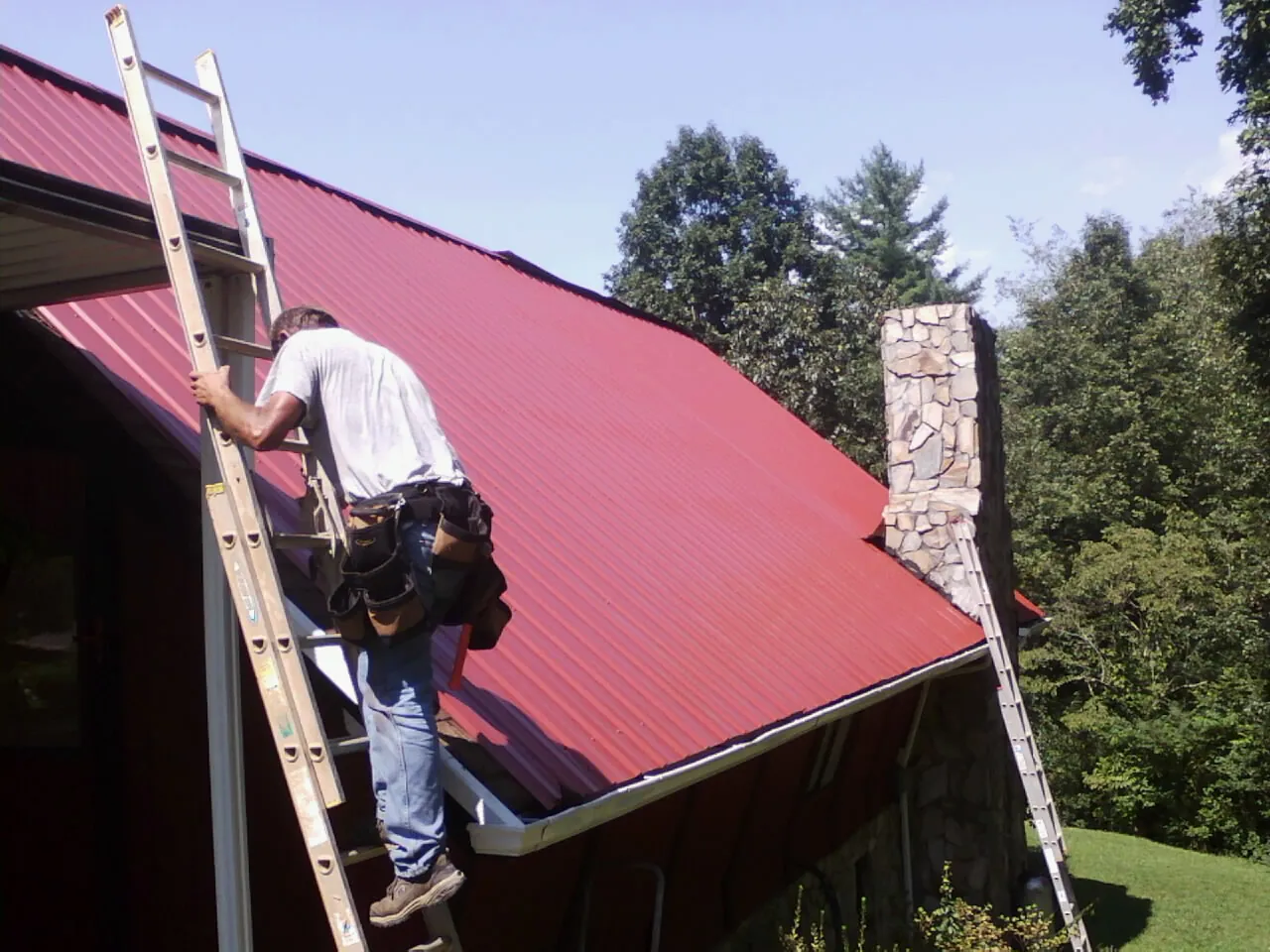 Roofer on a ladder beside a red metal roof and stone chimney — Soffit & Fascia Repair by Roofing Brothers in Lind